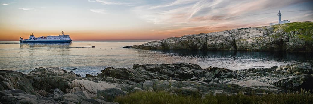 A large ferry sails near rocky shorelines at sunset, with a lighthouse visible on a grassy hill to the right and a colorful sky reflected on calm water.
